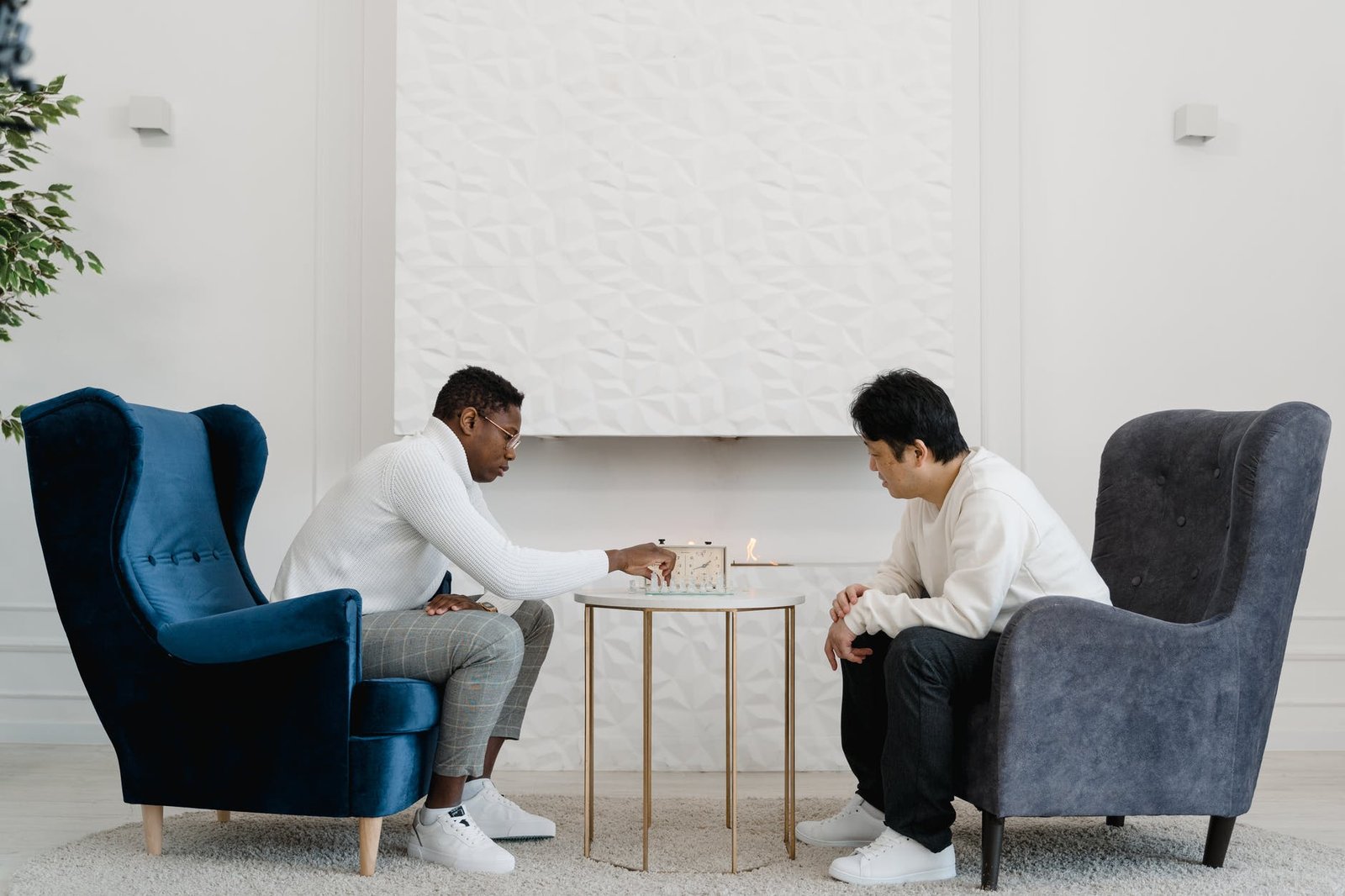 two men playing chess while sitting in armchairs beside white wall